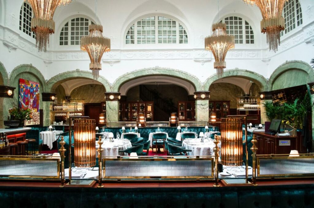Restaurante do Bank Hotel, em Estocolmo.O ambiente tem uma arquitetura histórica, com lustres, mesas com cadeiras estofadas, vasos com flores e obras e arte.