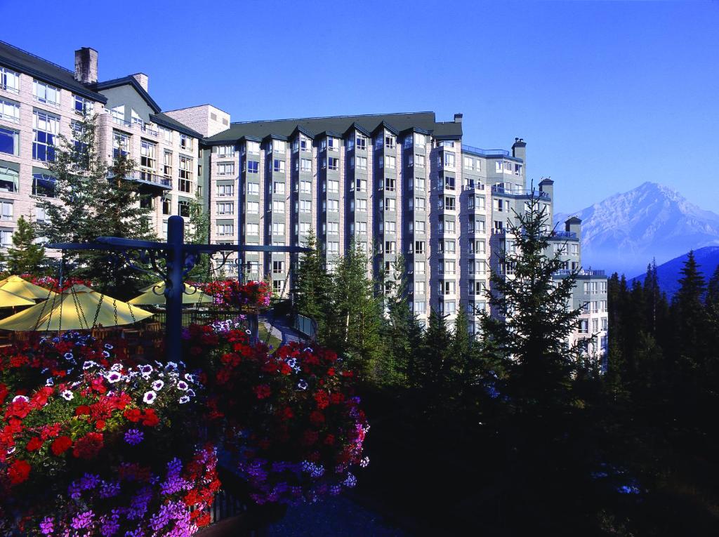 Imagem da parte de fora do Rimrock Resort Hotel que mostra a construção do hotel com vários andares e várias árvores e flores em volta durante o dia, ilustrando post Hotéis em Banff.