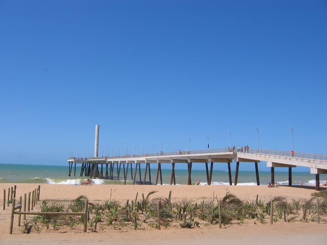 Grande píer de Costazul em Rio das Ostras que começa na areia e vai até o mar, em cima tem algumas pessoas andando e na faixa de areia tem outras pessoas. Imagem tirada durante o dia, ilustrando post Pousadas em Rio das Ostras.