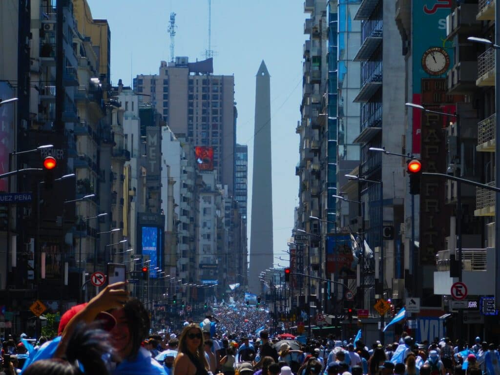 Rua com multidão de pessoas e prédios nos arredores, com o obelisco de fundo.