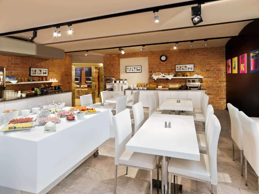Sala de café do Ibis Styles Stockholm Odenplan com mesas de cadeiras a frente e do lado esquerdo da imagem mesa com frios, frutas e aperitivos para o café da manhã.