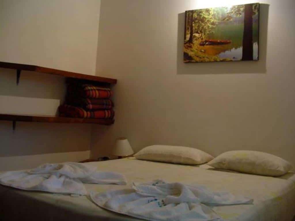 Quarto do Samburá Chalés. Uma cama de casal no meio, no lado esquerdo um abajur, duas prateleiras e cobertores em cima. Foto para ilustrar post sobre pousadas em Ilha Comprida.