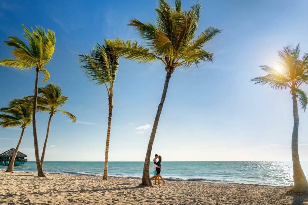 Casal dançando ao lado de uma palmeira na praia Juanillo. O sol brilha ao lado de outra árvore ali, e há mais delas ao redor.