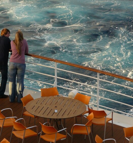Homem e mulher lado a lado encostados na grade de um navio enquanto olham para o mar em frente. Mesas e cadeiras estão espalhadas no canto inferior da imagem.