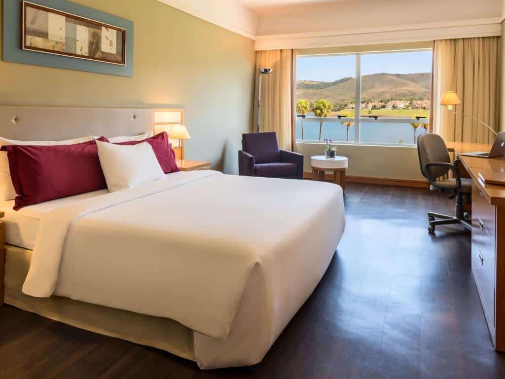 Suíte no Transamerica Lagoa dos Ingleses, representando o post sobre pousadas em Nova Lima. A cama é larga, box e de casal, e na foto ela está no canto inferior esquerdo. Do seu lado direito, há uma mesa de cabeceira com abajur e um vaso com flores. Mais a frente, há uma poltrona do lado de uma pequena mesa circular. No canto direito, vemos parcialmente uma cadeira giratória e uma escrivaninha com outro abajur. No fundo do quarto, estão as janelas com cortinas beges.