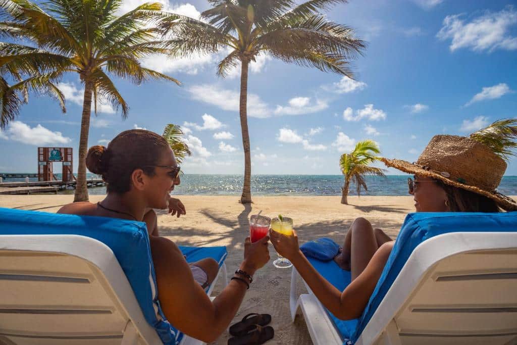 Foto de um homem e uma mulher sentados em espreguiçadeiras na beira da praia. Eles brindam com drinks enquanto conversam entre si. Na frente, podemos ver o mar e alguns coqueiros.