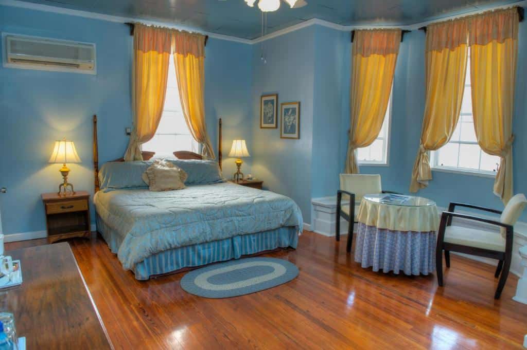 Quarto no The Great House Inn. A decoração é clássica e possui muito azul. A cama está no centro, encostada em uma parede. Atrás dela, há uma janela com cortinas amarelas. Dos lados da cama há uma cabeceira com abajur em cima. Do lado direito do quarto, há uma mesa redonda de vidro com duas poltronas em sua volta. Atrás há mais janelas. O piso é de madeira e as paredes são azuis.