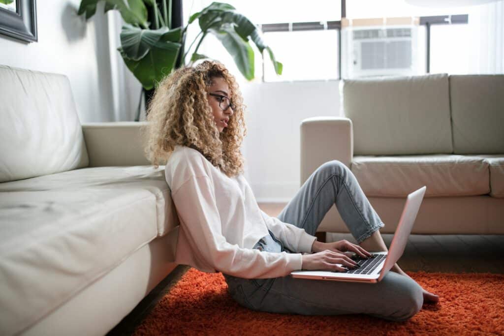 Uma mulher de calça jeans, camiseta branca e os cabelos enrolados usando um óculos, ela está sentada em um tapete e com as costas em um sofá, no colo dela há um notebook
