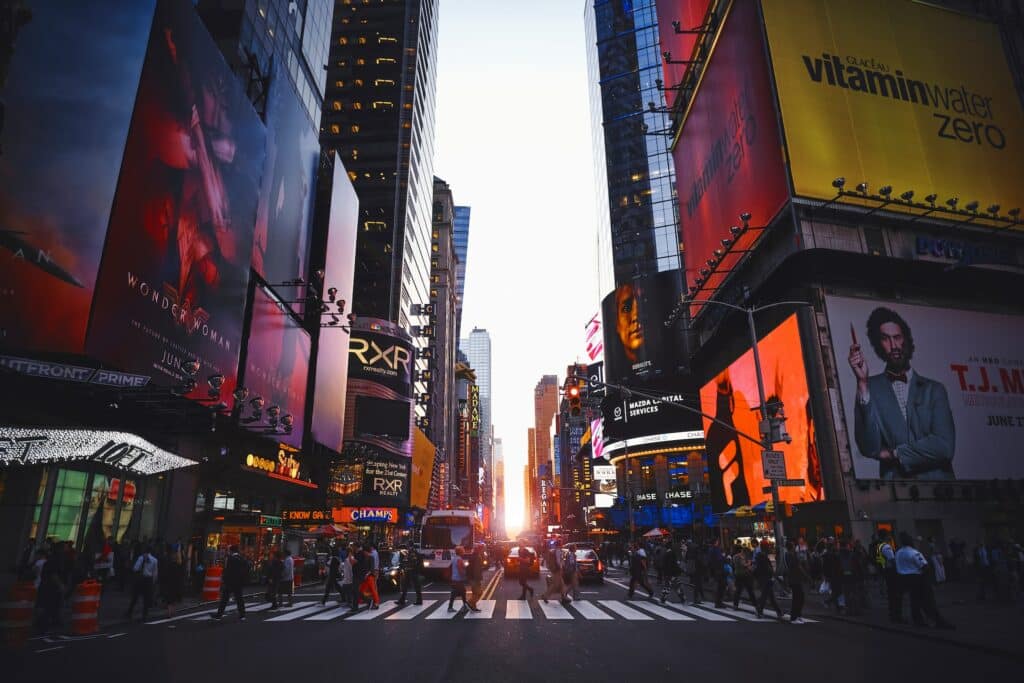 Vista ao entardecer na Times Square, uma das opções de o que fazer em Nova York, com várias pessoas passando por uma faixa de pedestres, enquanto telões enormes anunciam produtos eventos coloridos