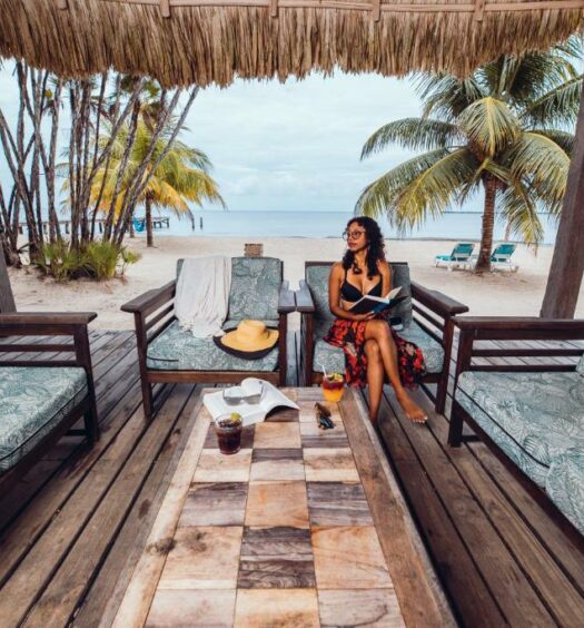 Uma mulher sentada em um sofá debaixo de um quiosque no Umaya resorts & Adventures, em Belize. O quiosque está sobre a areia e possui mais sofás e uma mesa no centro. A mulher segura um livro. Atrás dela, é possível ver o mar e alguns coqueiros.