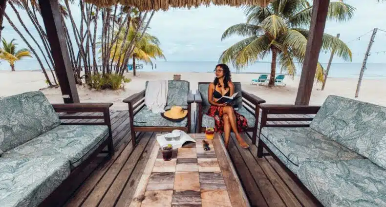 Uma mulher sentada em um sofá debaixo de um quiosque no Umaya resorts & Adventures, em Belize. O quiosque está sobre a areia e possui mais sofás e uma mesa no centro. A mulher segura um livro. Atrás dela, é possível ver o mar e alguns coqueiros. Representa o post de hotéis em Belize.