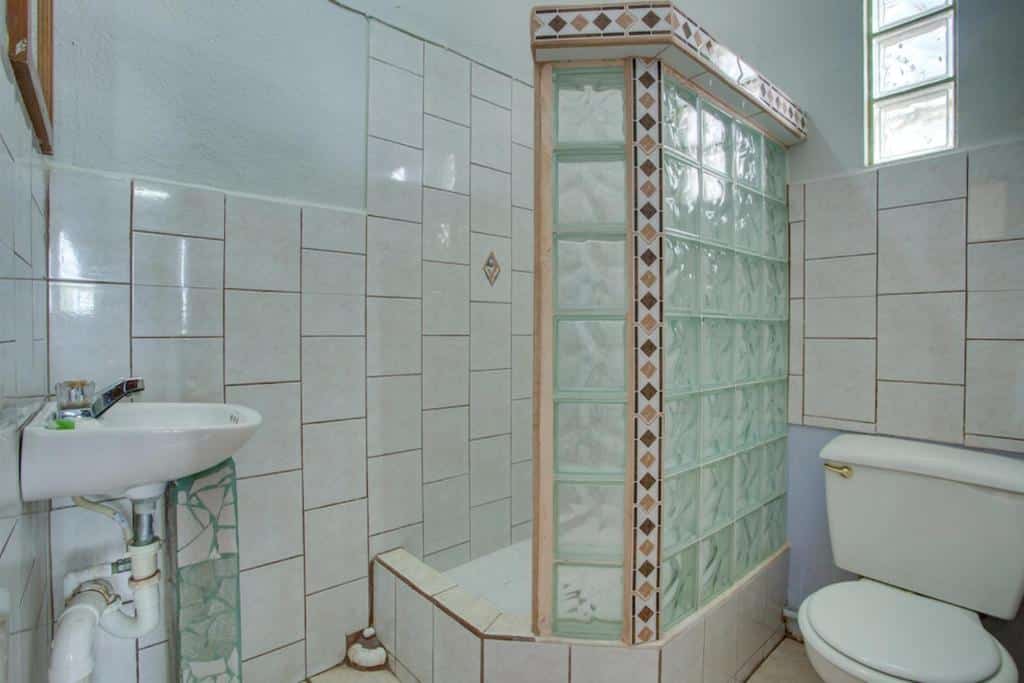 Banheiro de um quarto do Vênus Hotel. No canto inferior direito há uma privada. Do seu lado esquerdo há uma divisória de azulejos e vidro que separa a área da ducha. No canto inferior esquerdo está a pia.