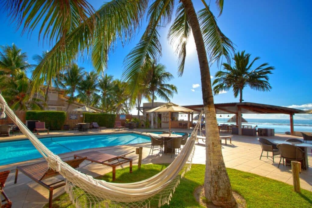 Rede pendurada em um coqueiro, com uma piscina atrás, espreguiçadeiras e mesas com guarda-sol espalhados em volta e o mar ao fundo do Village Barra Hotel, perto da praia do Gunga