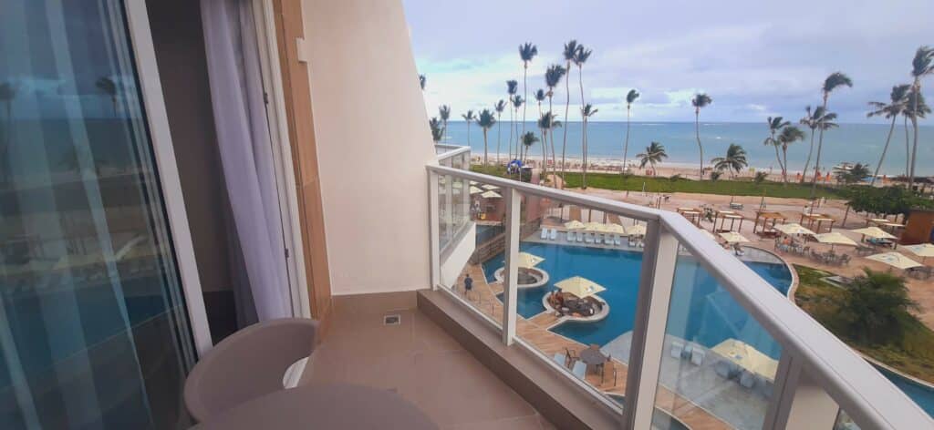 Varanda de um quarto do Maragogi Brisa Exclusive Hotel, com uma cadeira, mesa e vista para uma área de pisicnas, e a praia de Antunes com coqueiros balançando com o vento em Alagoas