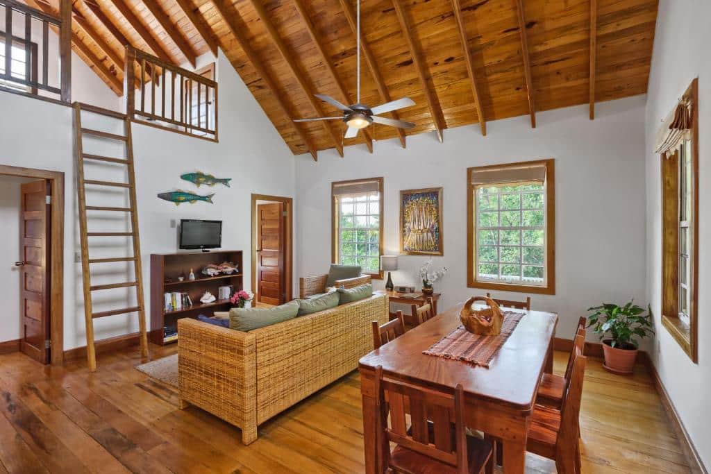 Imagem do interior do Weezie's Ocean Front Hotel and Garden Cottages. Temos uma visão da sala de estar e da área de jantar. Mais para a direita, há uma mesa de madeira com seis cadeiras, e na sua frente, o sofá. Na frente do sofá está uma pequena estante com prateleiras e uma TV. A cabana ainda possui um loft no segundo andar, com acesso por uma escada de madeira, essa que está do lado da estante da televisão. O lugar é espaçoso e possui janelas com entrada de luz natural.