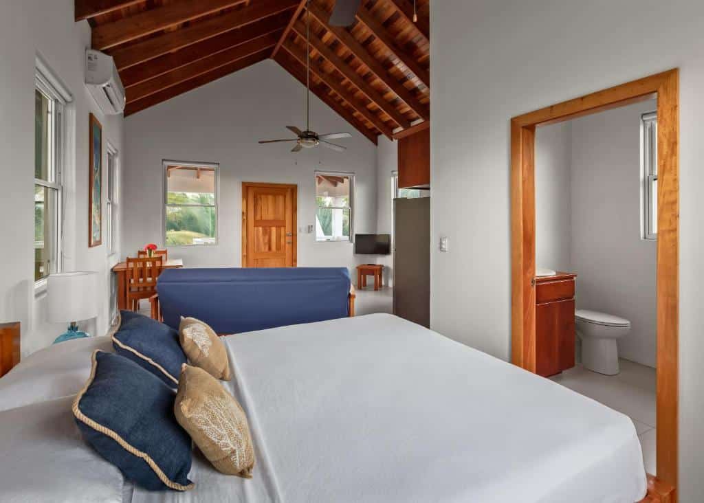 Imagem do Weezie's Ocean Front Hotel and Garden Cottages, para ilustrar o post sobre hotéis em Belize. A cama está em primeiro plano, na sua frente, no lado direito da foto, está a porta para o banheiro No fundo da imagem vemos um sofá, mesa com duas cadeiras, TV, a porta de acesso da cabana e parte da cozinha.