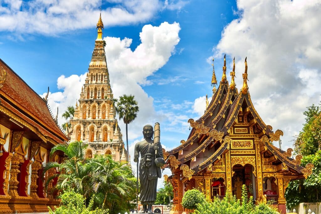 vista do templo budista Wiang Kum Kam, na Tailândia, com uma estátua de Buda na frente, além de torres atrás com arquitetura clássica do local, com muitos detalhes e cores vibrantes em dourado