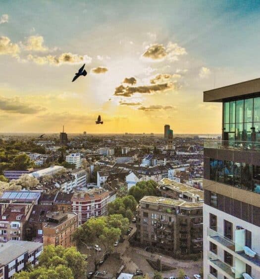 Vista aérea de Dusseldorf, com prédios e casas no horizonte e um céu com o pôr do sol refletindo em pássaros voando. À direita tem o prédio do 25hours Hotel Das Tour, um dos hoteis em Dusseldorf, com um letreiro escrito "25 hours"