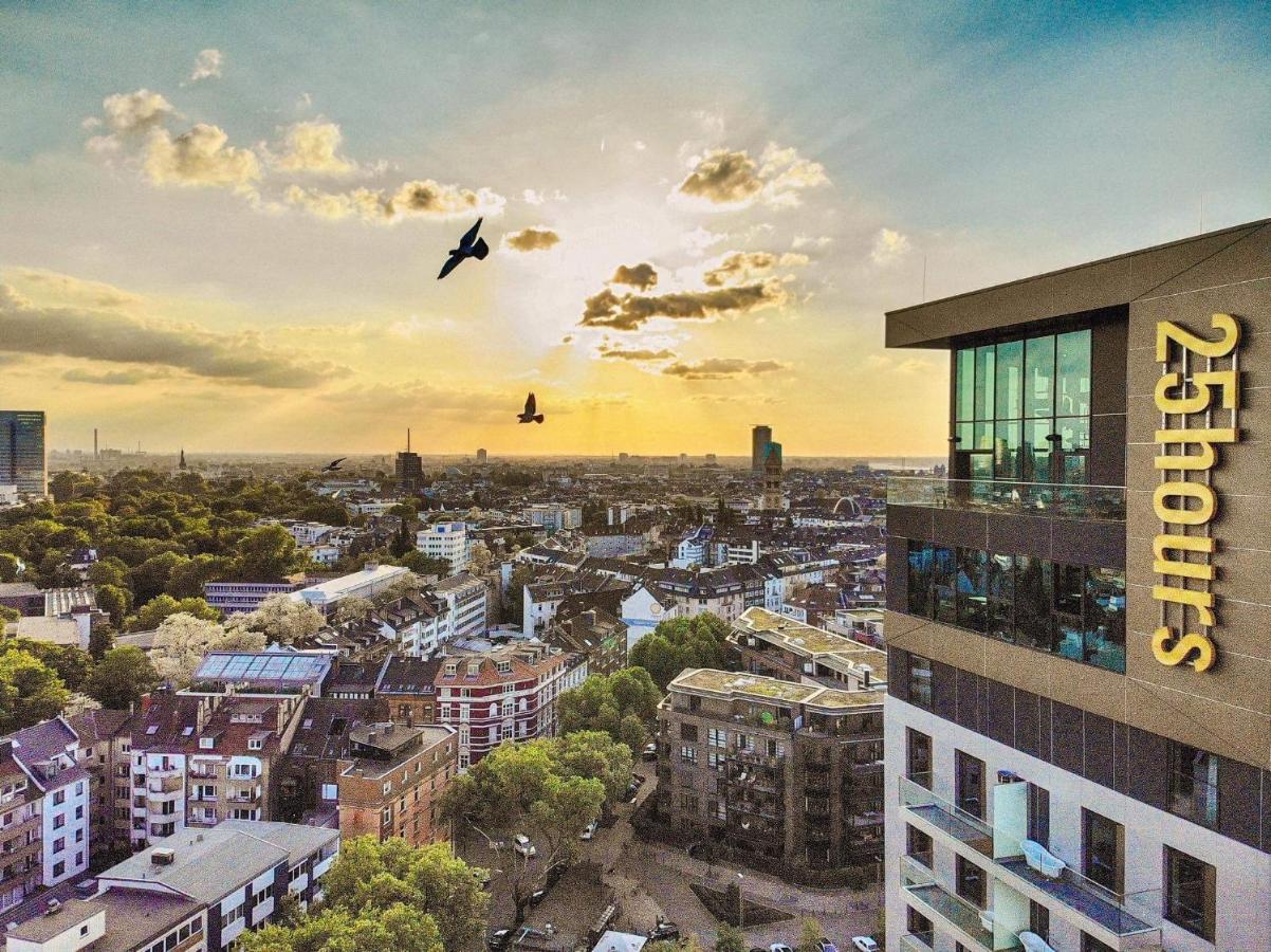 Vista aérea de Dusseldorf, com prédios e casas no horizonte e um céu com o pôr do sol refletindo em pássaros voando. À direita tem o prédio do 25hours Hotel Das Tour, um dos hoteis em Dusseldorf, com um letreiro escrito "25 hours"
