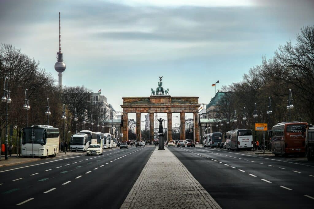 vista da rua com vários ônibus de ambos os lados com árvores e uma passarela no meio que chega até o Portão de Brandemburgo, em Berlim, com uma escultura de anjos em cima