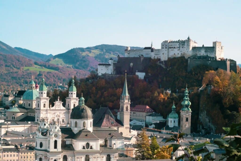 vista da cidade de Salzburg, na Áustria, com um castelo ao fundo, paisagens montanhosas e verdejantes, além de prédios e mais casinhas típicas