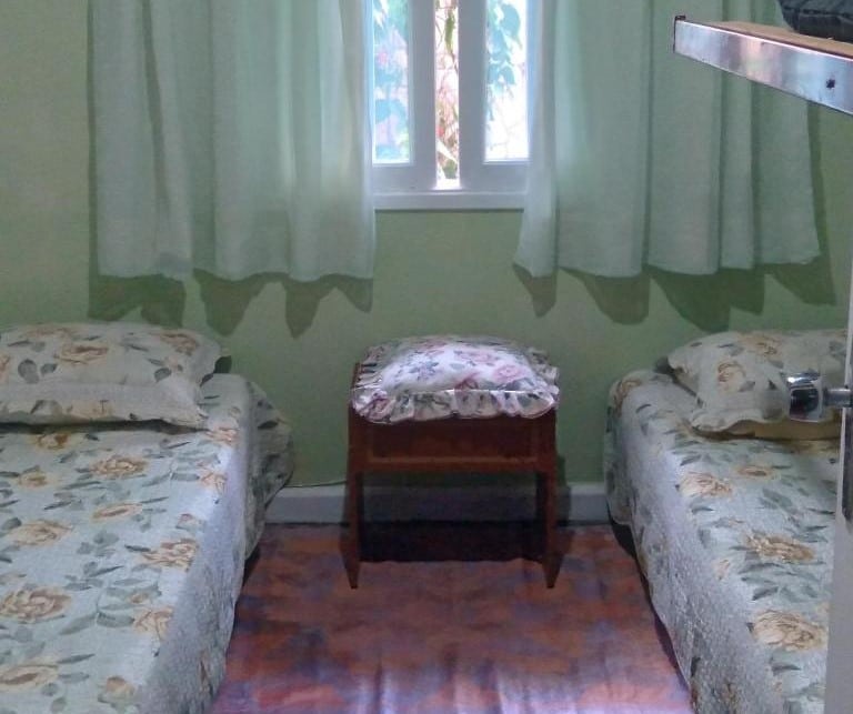 Quarto da A Casa do Chef Ubatuba. Uma cama de solteiro de cada lado, no centro um banco de madeira. Em cima uma janela com cortinas. Foto para ilustrar post sobre airbnb em Perequê Mirim.