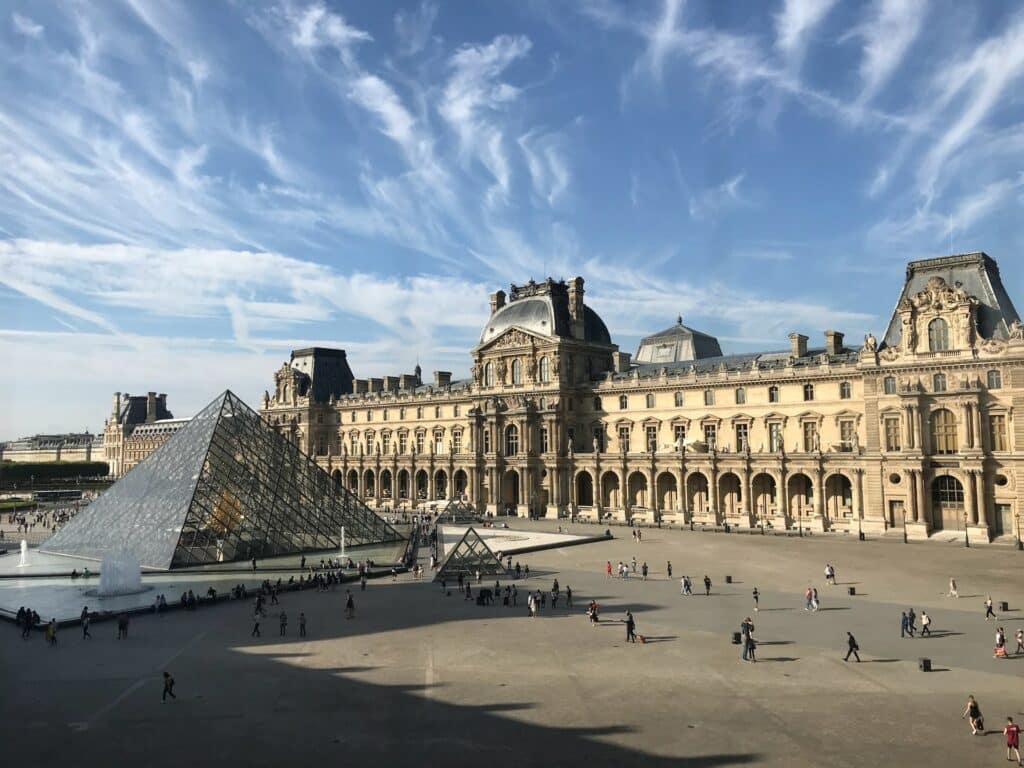 O Museu do Louvre, em Paris, é um extenso palácio de fachada com colunas. Logo em frente a ele e na lateral esquerda da imagem fica uma pirâmide alta de vidro. Várias pessoas passeiam ao redor durante o dia de céu azul. - Foto: Ahmad via Unsplash