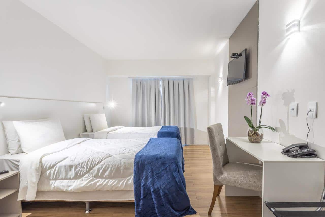 Quarto do Americas Townhouse Hotel. Duas camas de solteiro do lado esquerdo, de cada lado uma mesa de apoio e um lustre. Do lado direito uma mesa de trabalho e uma TV. No fundo a janela com a cortina fechada. Imagem para ilustrar post sobre hotéis perto do Rock in Rio.