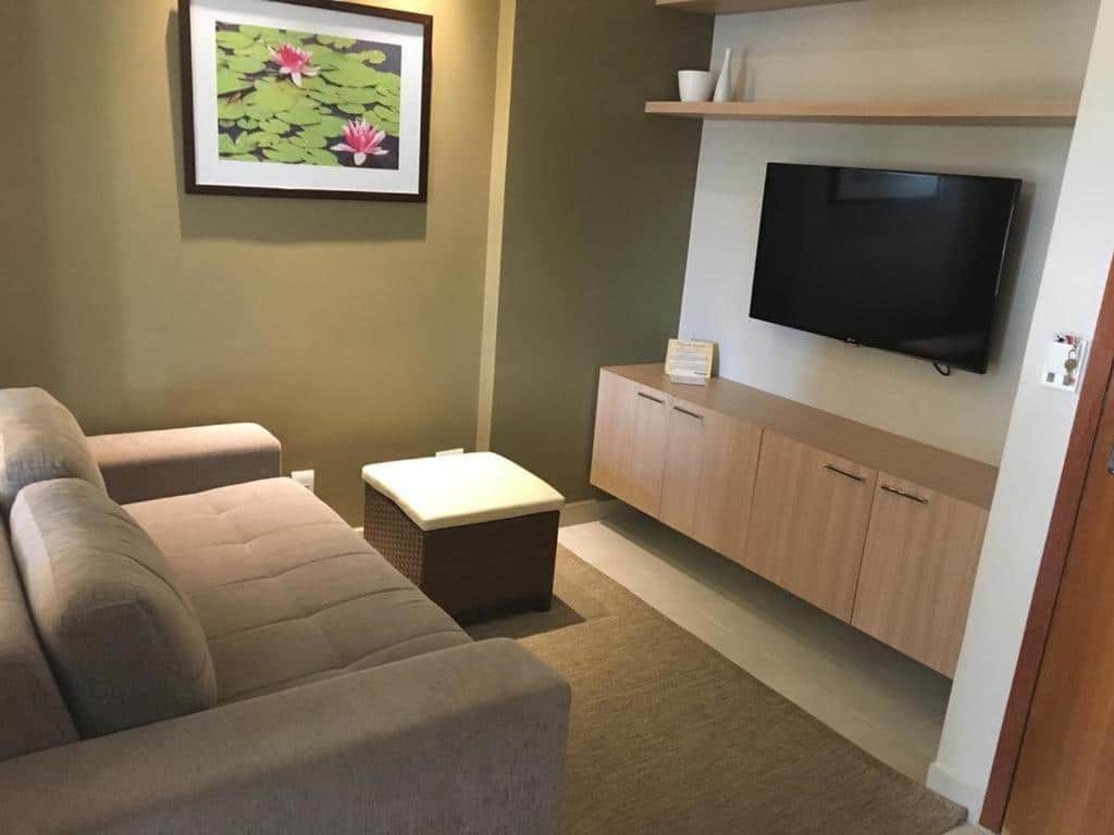 Apartamentos Caldas Novas. Do lado esquerdo um sofá, no meio um puff, do lado direito um armário e uma televisão em cima.
