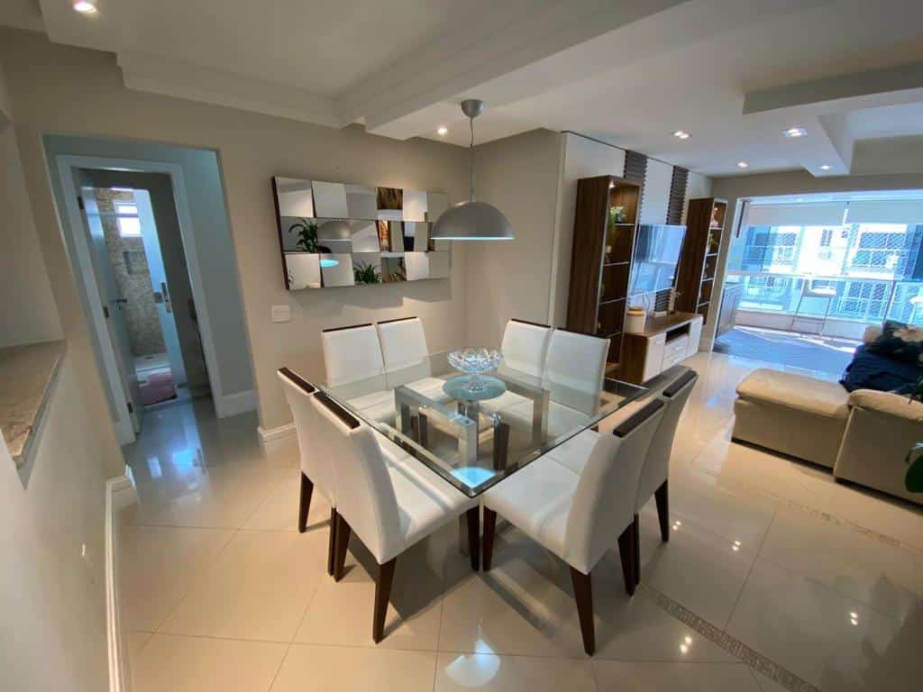 Sala de estar e cozinha do Apartamento com 3 dormitórios, em Balneário Camboriú. Do lado esquerdo existe uma mesa quadrada de oito lugares e, ao fundo, um corredor e uma porta. Do lado esquerdo está a sala, com um sofá, uma estante e uma tv.