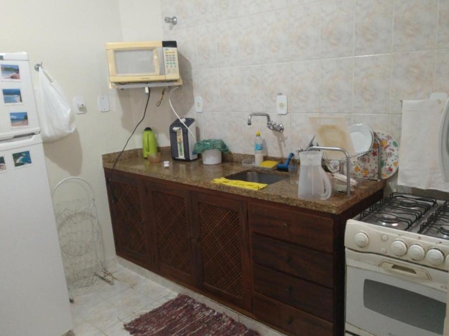 Cozinha do Apartamento em Ubatuba. Do lado esquerdo uma geladeira, e uma fruteira vazia. Do lado direito um fogão, uma pia com escorredor de louça, em cima um micro-ondas.