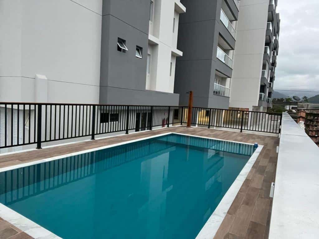 Área externa do Apartamento Jardins Ubatuba. Uma piscina ao ar livre com grade ao redor. No fundo os apartamentos.