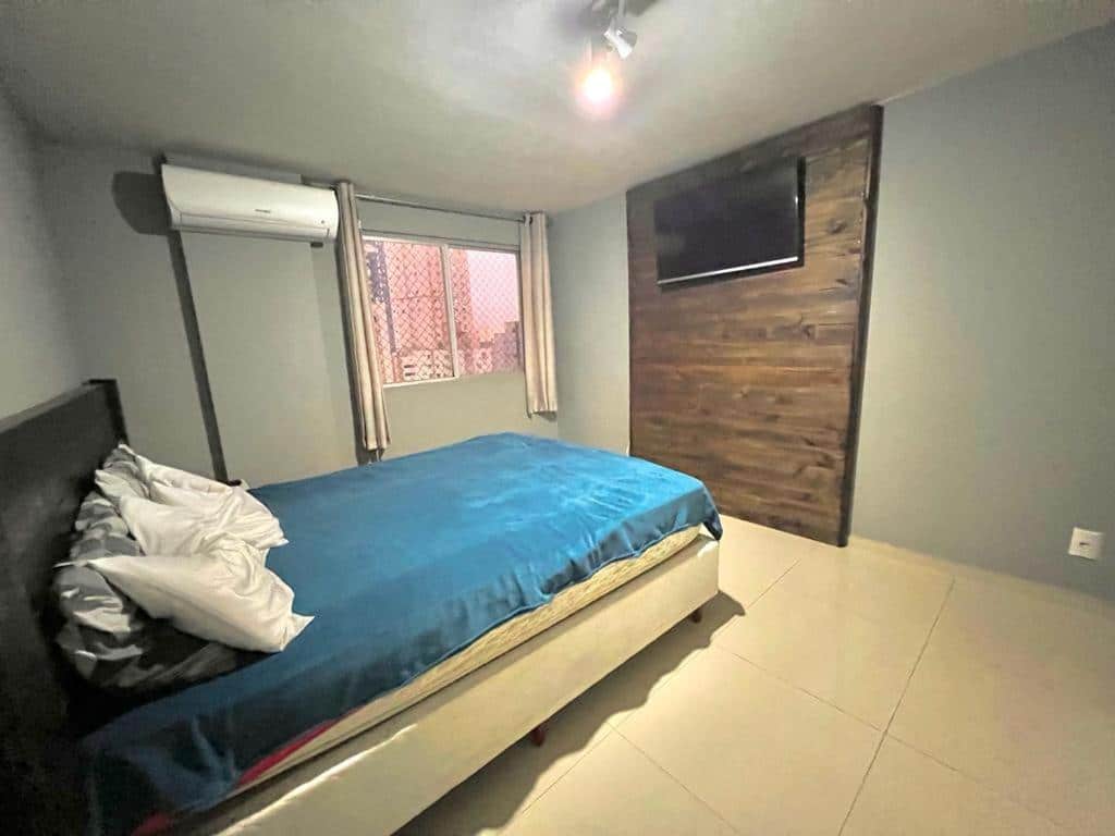 Quarto do Apartamento Top. Uma cama de casal e de frente uma televisão. No fundo, um ar-condicionado e uma janela. Foto para ilustrar post sobre airbnb em Balneário Camboriú.