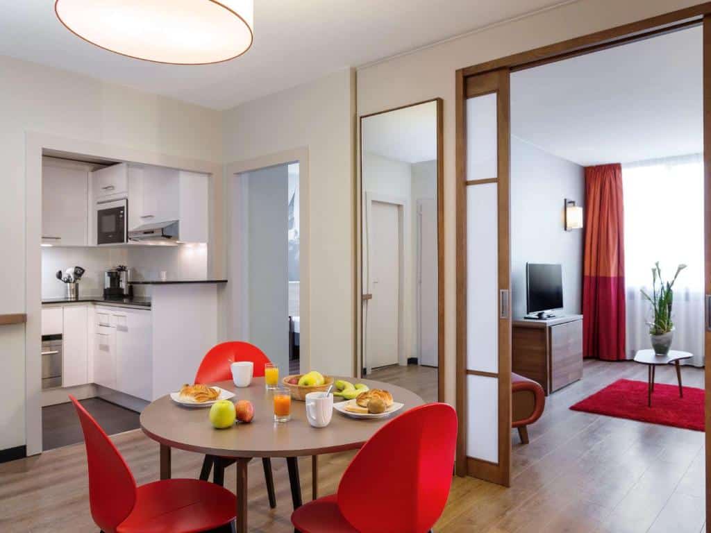 Imagem do interior de um apartamento do Aparthotel Adagio Genève Mont-Blanc. Em primeiro plano vemos uma mesa redonda de madeira com três cadeiras vermelhas. Em cima há pratos com café da manhã. Na esquerda, no fundo, há uma cozinha e um corredor para os quartos. Na direita, uma divisória que adentra a sala de etar.