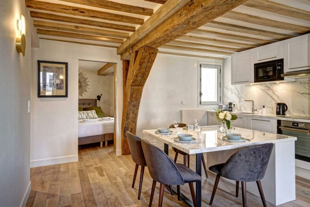 Interior de um dos Apartments Du Louvre St Honoré. A foto foi tirada na sala de jantar, que tem uma mesa posta com quatro cadeiras. Uma bancada branca separa o local da cozinha equipada com armários e fogão. Na parede ao fundo há uma janelinha. Na mesma parede da janela há uma porta que leva ao quarto, onde é possível ver uma cama de casal.