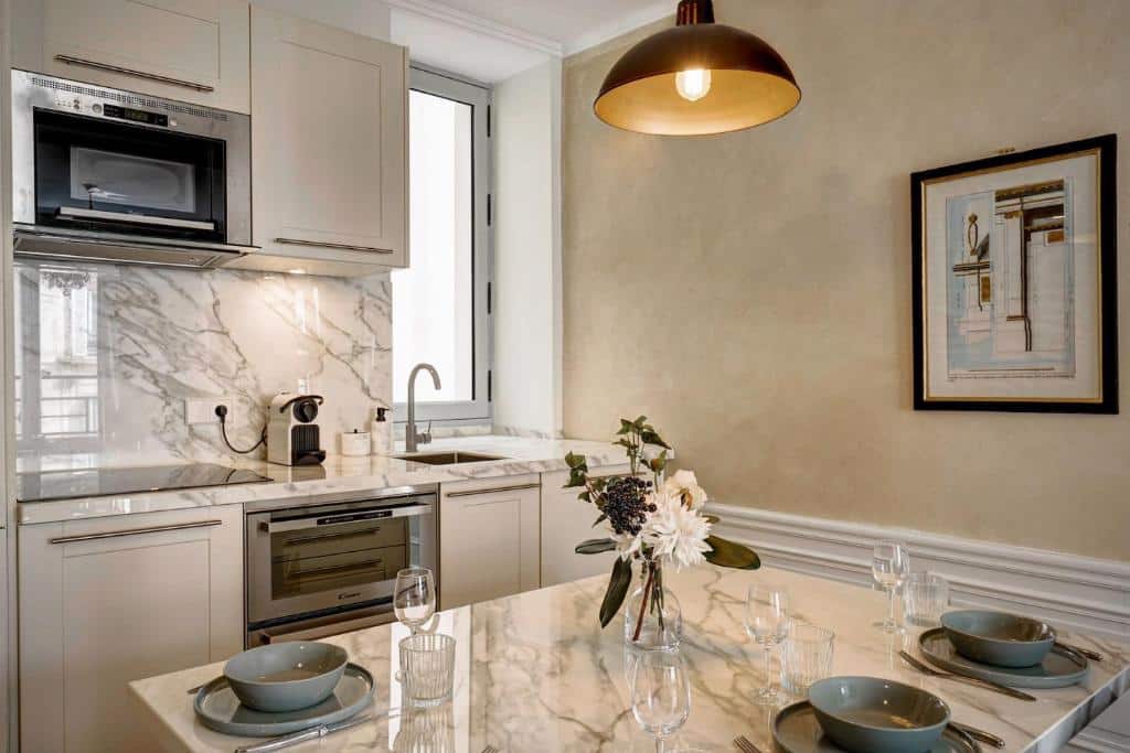 Cozinha dos Apartments Du Louvre St Honoré. Uma bancada de pedra tem pratos e taças e, ao fundo, bancadas e armários contam com forno, cooktop, cafeteira, pia e micro-ondas. Há uma janela em frente à pia.