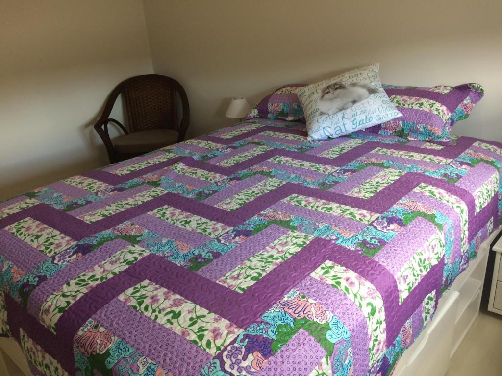 Quarto do Serenidade em Canela. Há uma cama de casal com roupas de cama colorida e um travesseiro com desenho de gato estampado. No seu lado esquerdo há um abajur e uma cadeira. Representa o post sobre airbnb em Canela.