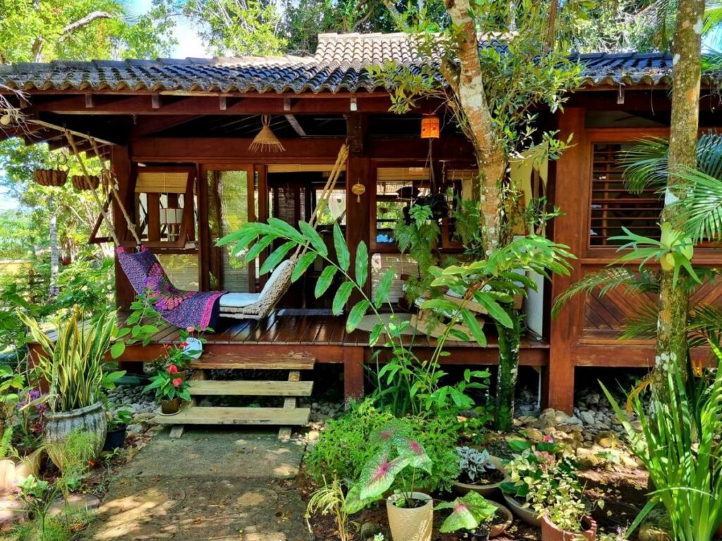 Entrada do Bangalô alto do Mucugê. Ele é todo de madeira, possui uma pequena escadinha para entrar e uma varanda com uma rede. Ele está cercado por árvores e plantas. Esse é um airbnb em Arraial d'Ajuda.