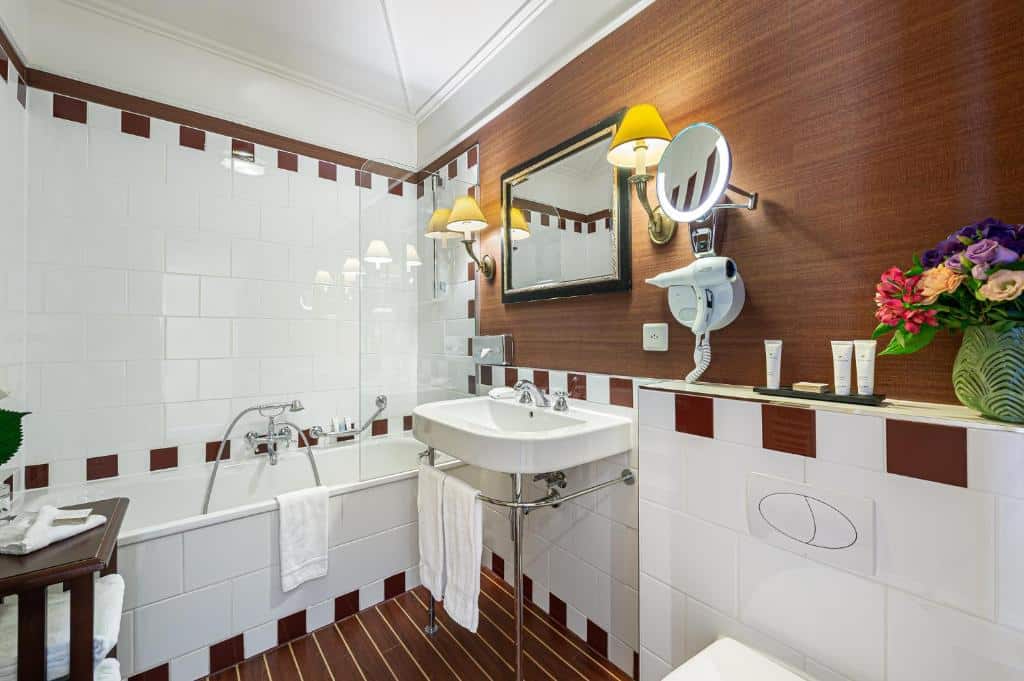 Banheiro do Kipling Manotel. Há um pia com barras, um espelho, dois abajures, e secador de cabelo. Na esquerda ada pia, encostada na parede, há uma banheira de azulejos com ducha e barras de apoio. J´no canto inferior direito, vemos de relance a privada e uma bancada com amenidades e um vaso de flor em cima.