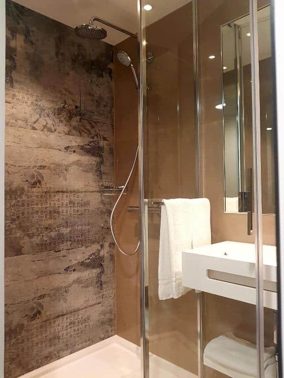 Imagem do banheiro do Nash Pratik Hotel As paredes são marrons e a do box possui um papel de parede no mesmo tom. No canto inferior direito há uma pia branca e espelho.