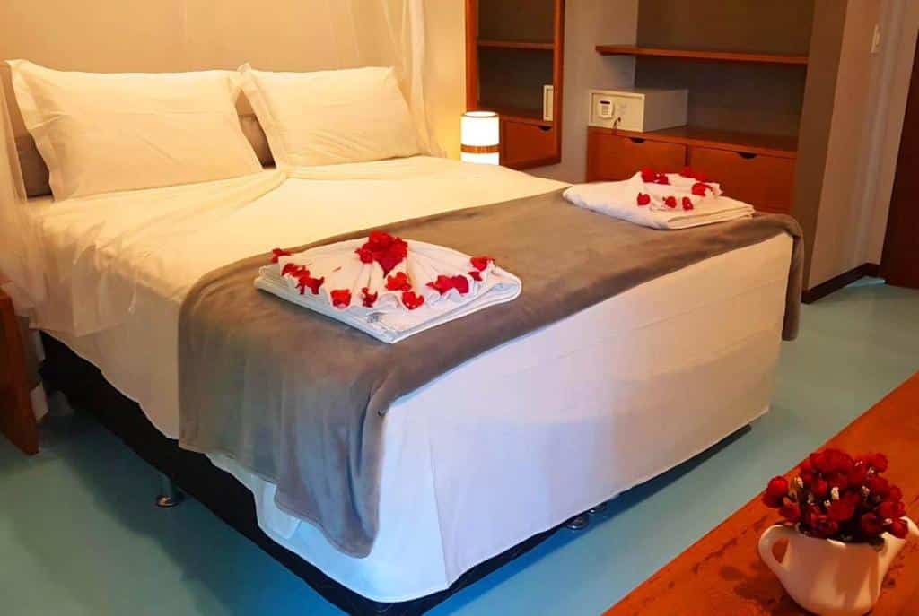 Um dos quartos do BELLA VISTA Village. Uma cama de casal está no meio, com toalhas cheias de pétalas de rosa. Ao seu lado direito há um armário com um cofre.