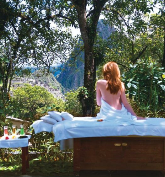 mulher branca de cabelos ruivos sentada em cima de uma maca de massagem com as costas nuas, olhando para as ruínas da cidade de Machupicchu ao fundo, escondida pela vegetação verde ao redor
