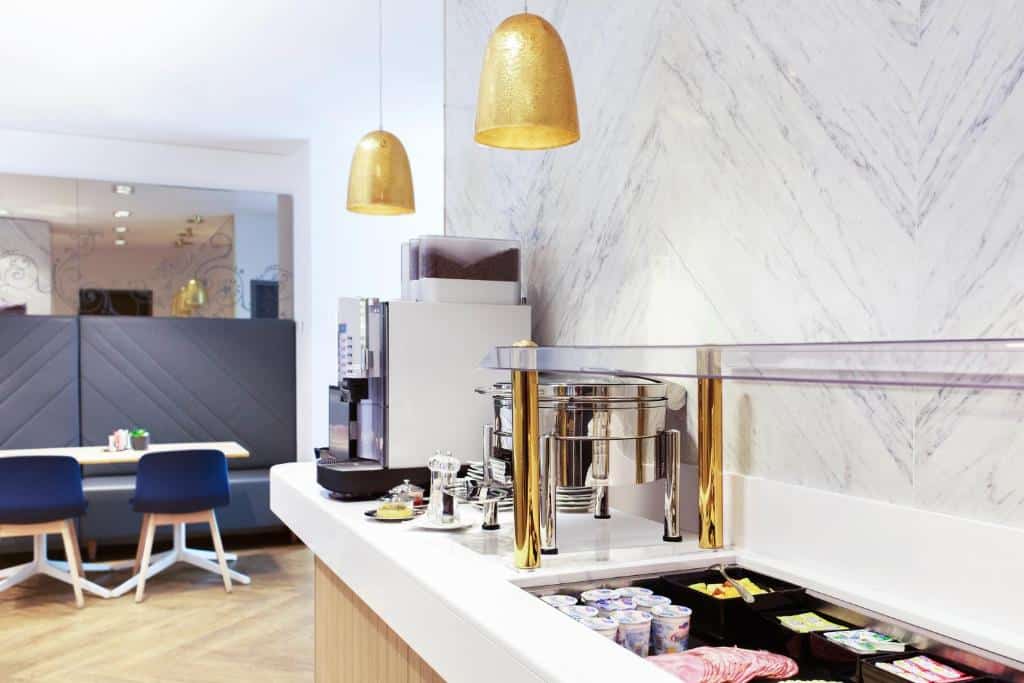 Restaurante no VISIONAPARTMENTS Geneva Gare. A bancada com buffet está na direita, com opções de iogurte, frios, etc. Há também uma máquina de café. No fundo, no lado esquerdo, vemos as mesas com cadeiras.