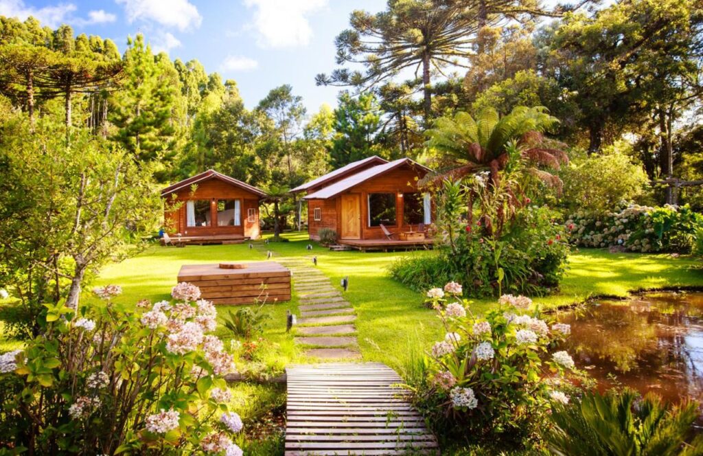 Frente das Cabanas Parque Suíço. Uma ponto no jardim e atrás duas cabanas com jardim ao redor.