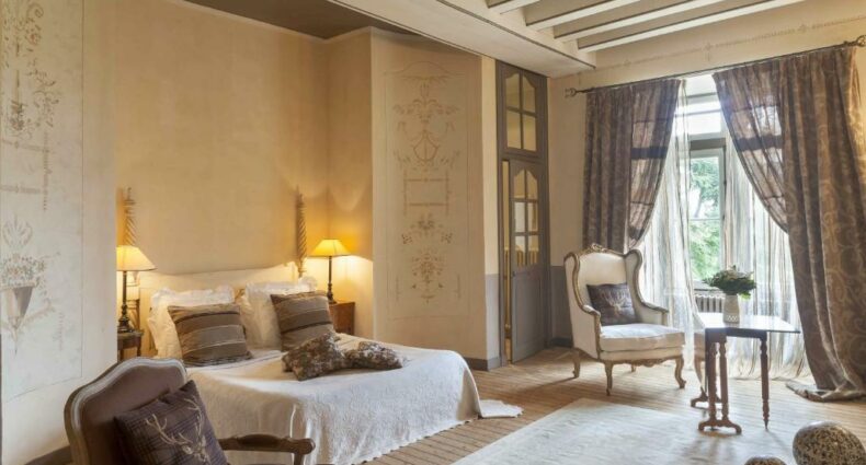 Quarto do Château de l'Epinay, com cama de casal do lado esquerdo da imagem em cada lado da cama uma comoda com luminária, do lado esquerdo uma poltrona com uma mesinha. Representa hotéis no Vale do Loire.