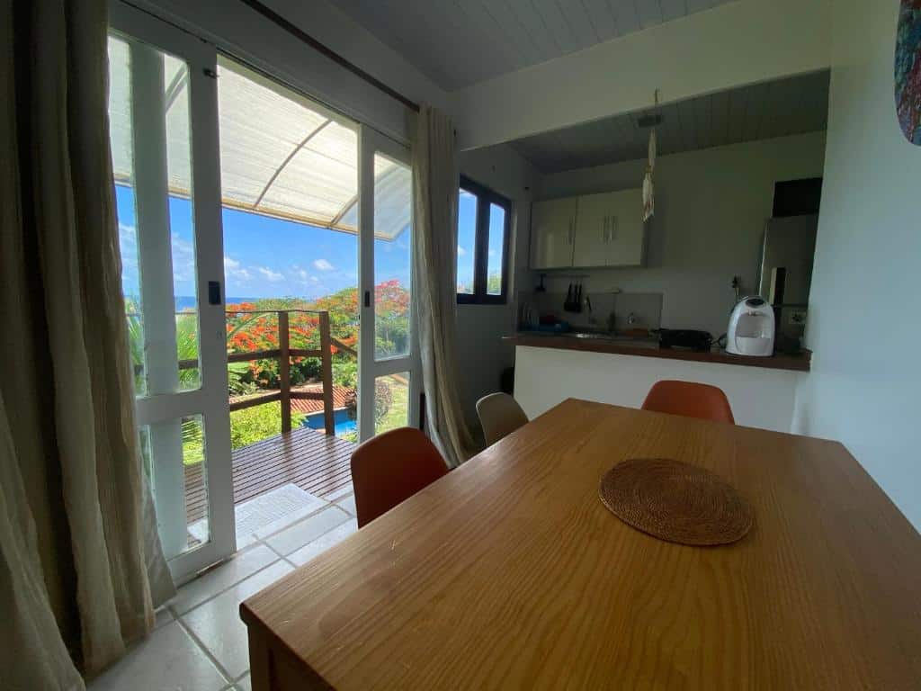 Cozinha da Casa do Ney. Uma mesa do lado direito, no fundo um balcão com a cozinha, do lado esquerdo uma porta de vidro com vista para o mar e a varanda.