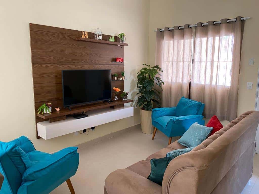 Sala de estar do Casa em Brotas, Refúgio e Aconchego com uma sofá, duas poltronas, um vaso de planta no canto direito perto da janela com cortina e no centro da parede tem uma estante de madeira com uma tv.