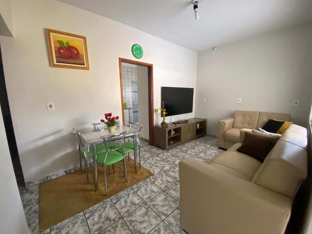 Sala de estar da Casa em Caldas Novas. Do lado direito dois sofás, de frente uma raque com televisão, uma mesa de vidro de quatro cadeiras e a porta da cozinha.
