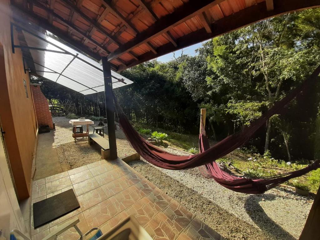 Área externa da Casa Kiiro. Duas redes do lado direito com vista para o jardim, do ado esquerdo, no fundo, uma churrasqueira e uma mesa de madeira com bancos.