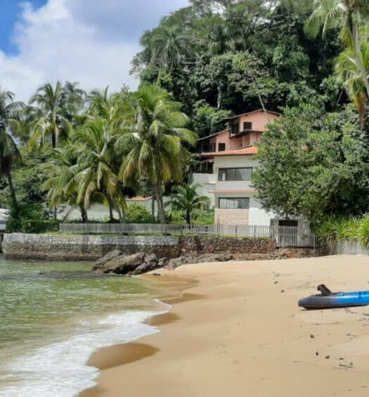 Casa de temporada vista da praia. Em volta da casa há uma vegetação com árvores e coqueiros e no lado esquerdo está o mar. Imagem para ilustrar o post airbnb em Angra dos Reis.